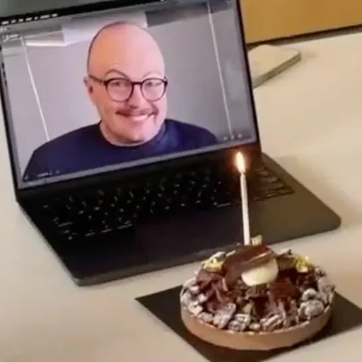 Kapwing marketing image of a virtual birthday celebration, featuring a cake with a lit candle in front of a laptop displaying a smiling man on a video call.