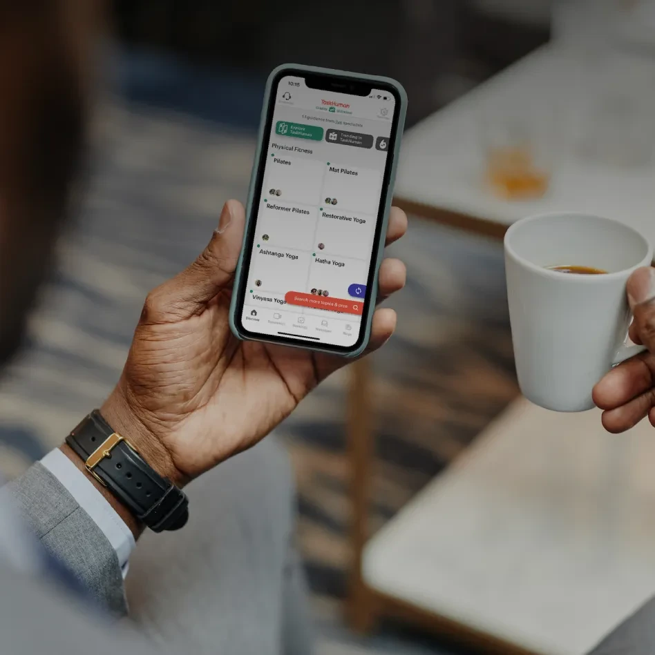 TaskHuman mobile app interface displaying various physical fitness categories like Pilates and Yoga, held by a person also holding a coffee cup.
