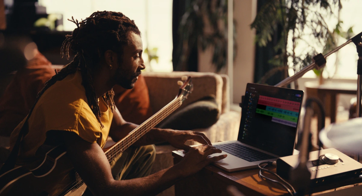 Suno user producing music in a home studio, playing bass guitar and interacting with a digital audio workstation on a laptop.